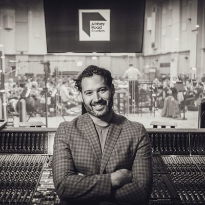 Lucas Cantor Santiago stands smiling with arms crossed in front of a large audio mixing console at Abbey Road Studios, with an orchestra visible through the glass window behind him.