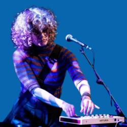 A musician with curly hair performing on stage, playing a small keyboard with a microphone in front of her. She wears a striped top and stands against a blue backdrop, focused on her performance.