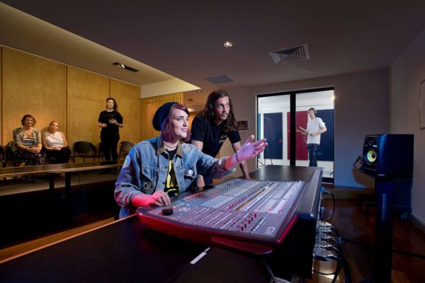 Two people sit at an audio mixing console in a recording studio, one pointing at controls. Three people observe from chairs in the background, while another stands behind glass in a recording booth.