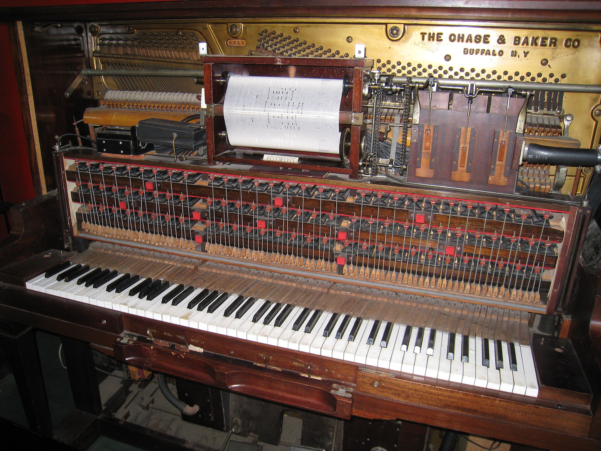 A close-up of a vintage player piano showing exposed keys, hammers, wires, and a paper music roll mechanism, labeled “The Chase & Baker Co., Buffalo, N.Y.” on a brass plate above the keyboard.