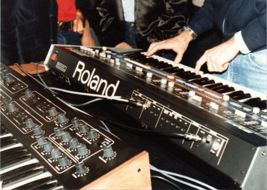 A person playing a Roland synthesizer keyboard, with another analog synthesizer featuring multiple knobs and sliders in the foreground. Several people stand nearby, some wearing jackets and jeans.