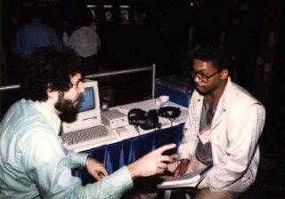 Paul Lehrman and Herbie Hancock  talking MIDI at a booth with a vintage computer and headphones on the table between them. One gestures while speaking; the other holds a notepad and listens attentively. The background is dimly lit with several people visible.