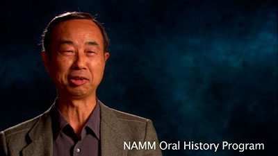 A man in a gray blazer smiles against a dark blue background. The text NAMM Oral History Program appears in the lower right corner.