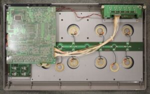 An open arcade controller reveals its internal components, including circuit boards, wiring, and eight round button mechanisms arranged in two rows.