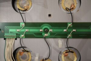 Close-up of an electronic circuit board with green traces, wires, and connectors, surrounded by four circular components with visible wiring, mounted on a gray surface.