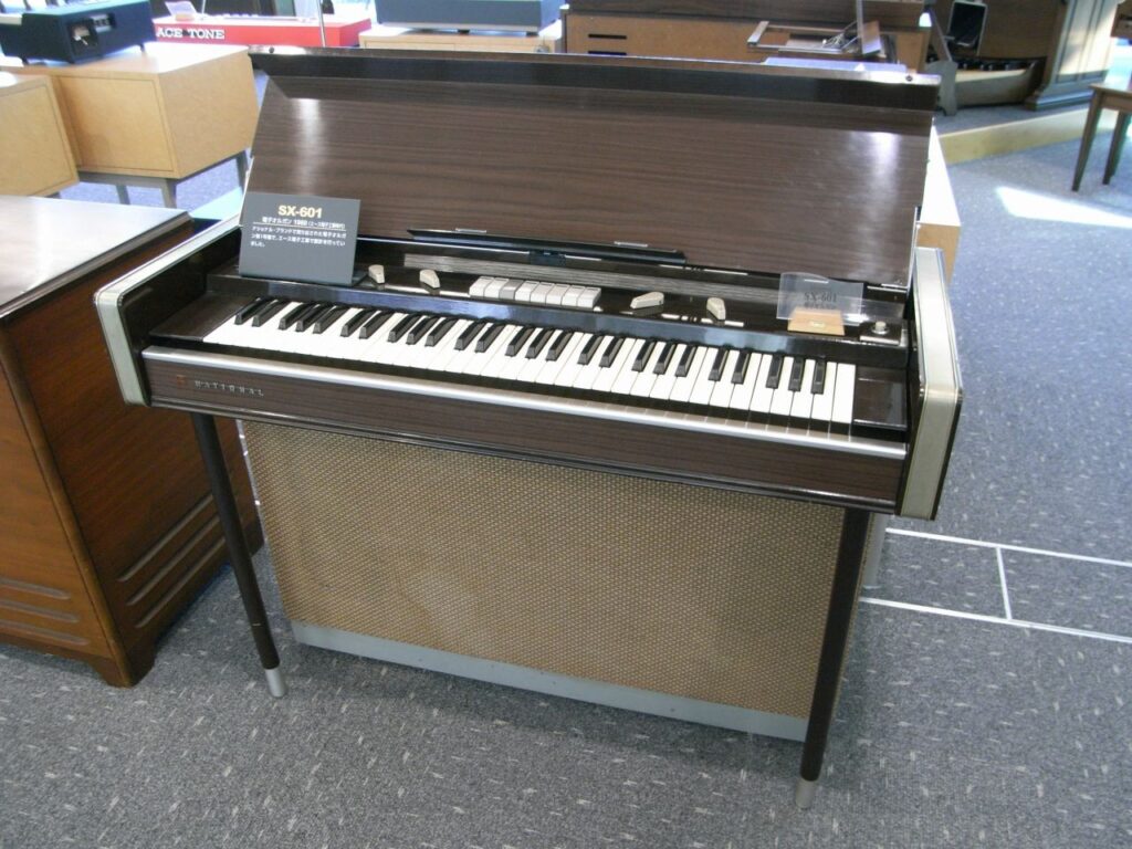 A vintage brown and beige electric organ with a raised lid, partially showing internal components, and white and black keys. It stands on four legs on a carpeted floor in a showroom with other instruments.