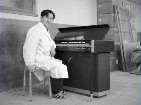 A man in a lab coat and sandals sits on a stool beside an open electronic organ in a sparsely furnished room, looking at the camera and smiling.