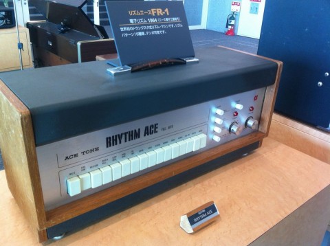A vintage Ace Tone Rhythm Ace drum machine with white switches, knobs, and a display sign on top, placed on a wooden table in a retro-styled room.