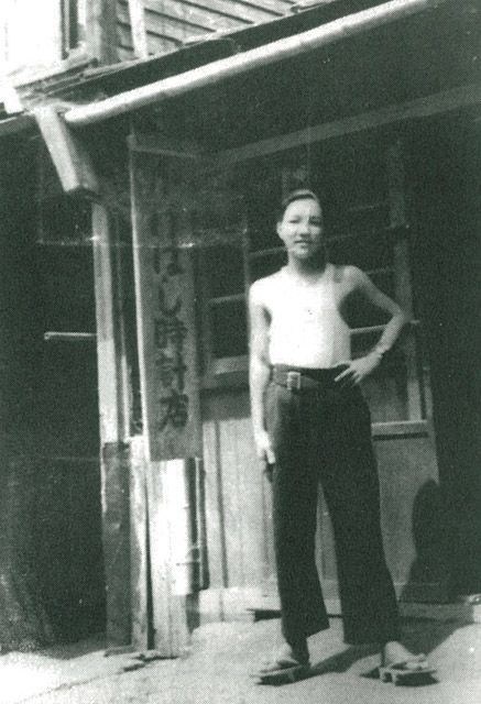 A man stands outside a wooden building, shirtless and wearing dark pants and sandals. He has one hand on his hip. A vertical sign with Japanese writing hangs near the entrance. The photo appears old and black-and-white.