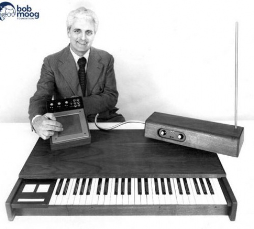 A man in a suit sits behind a keyboard instrument, holding an electronic device. Next to him is a theremin with an upright antenna. The image is black and white, with a Bob Moog Foundation logo in the corner.
