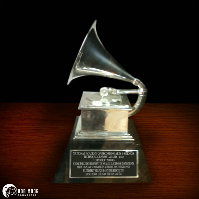 A silver Grammy Award trophy shaped like a gramophone sits on a wooden surface. The plaque on the front recognizes Dr. Robert Moog for his technical contributions to music. The Bob Moog Foundation logo is visible in the corner.