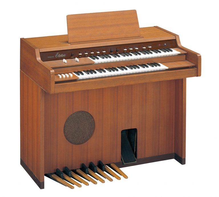 A vintage wooden electric organ with two keyboards, multiple control switches, a music stand, a speaker, and foot pedals at the base.