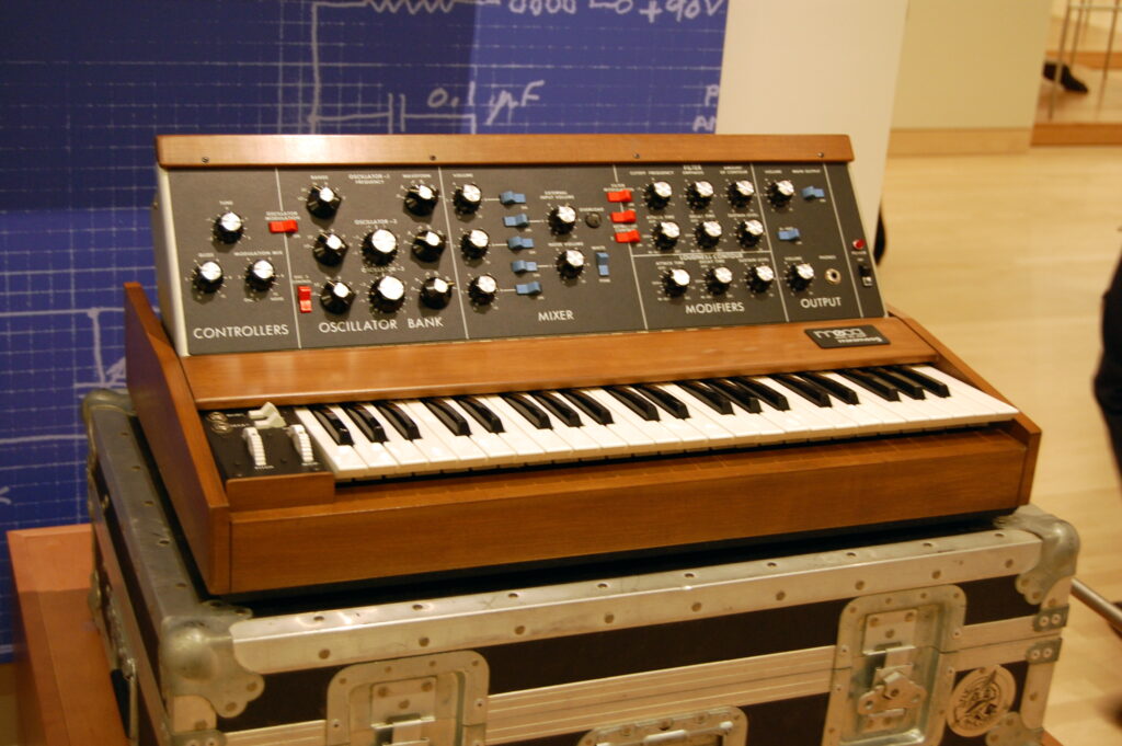 A vintage analog synthesizer with wooden side panels and many knobs sits on a metal equipment case, with a blue technical diagram in the background.