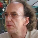 A middle-aged man with glasses and thinning, wavy brown hair looks to the left. He is wearing a light-colored shirt and appears to be outdoors, with blurred umbrellas and greenery in the background.