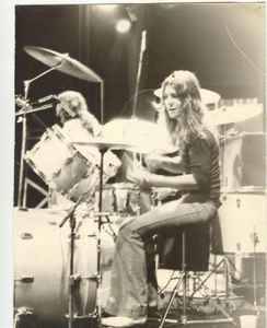 Black-and-white photo of a drummer with long hair playing a drum set on stage, wearing a dark shirt and jeans. Stage lights are visible in the background.