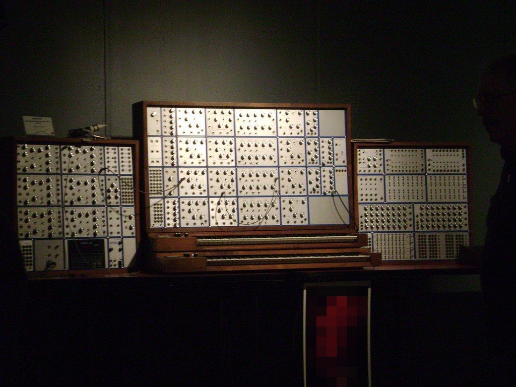 A large vintage modular synthesizer with many dials, switches, and cables is displayed in a dark room. The equipment is arranged in three wooden-framed panels on a table, with some wires visibly connecting modules.