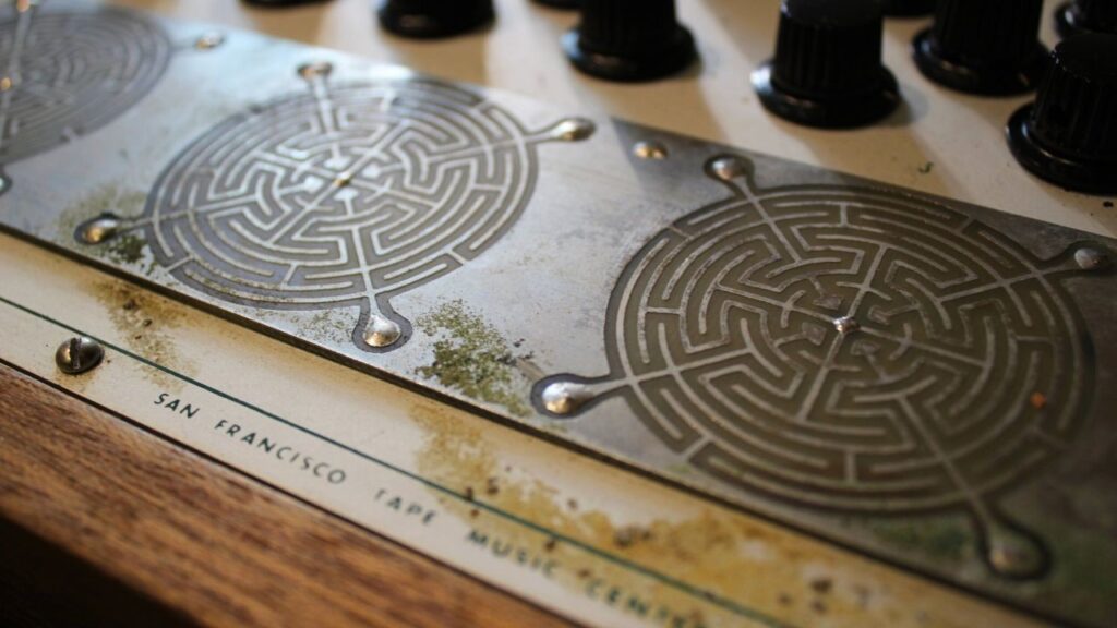 Close-up of a vintage electronic device with metal circular maze patterns, black knobs, and some grime. San Francisco and TAPE MUSIC are partially visible on the lower panel.