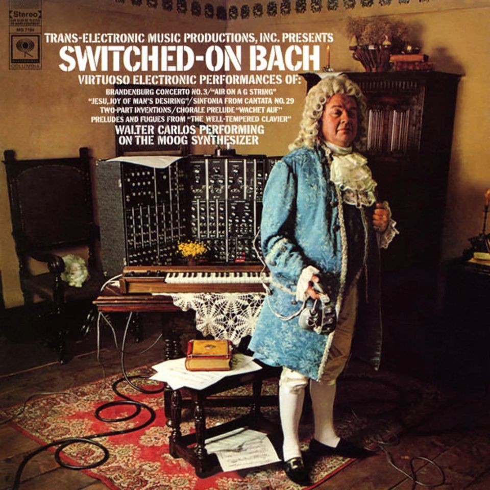 A person in 18th-century attire stands beside a large synthesizer in a room with a harpsichord and ornate rugs. The text above promotes Wendy Carloss Switched-On Bach album, featuring Bach music performed on a Moog synthesizer.
