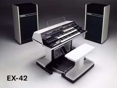 A vintage Yamaha EX-42 electronic organ with dual keyboards, foot pedals, a matching bench, and two large speakers, displayed in a studio setting. The text EX-42 appears in the lower left corner.