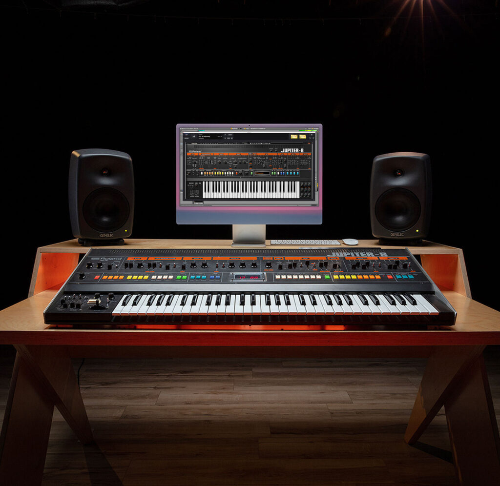 A modern music studio setup with a synthesizer keyboard, computer monitor displaying virtual synthesizer software, and two speakers on a wooden desk against a dark background.