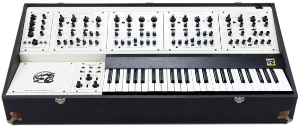 A vintage white and black analog synthesizer with multiple control knobs, switches, and sliders above a keyboard, all housed in a rectangular black case with metal corner protectors.