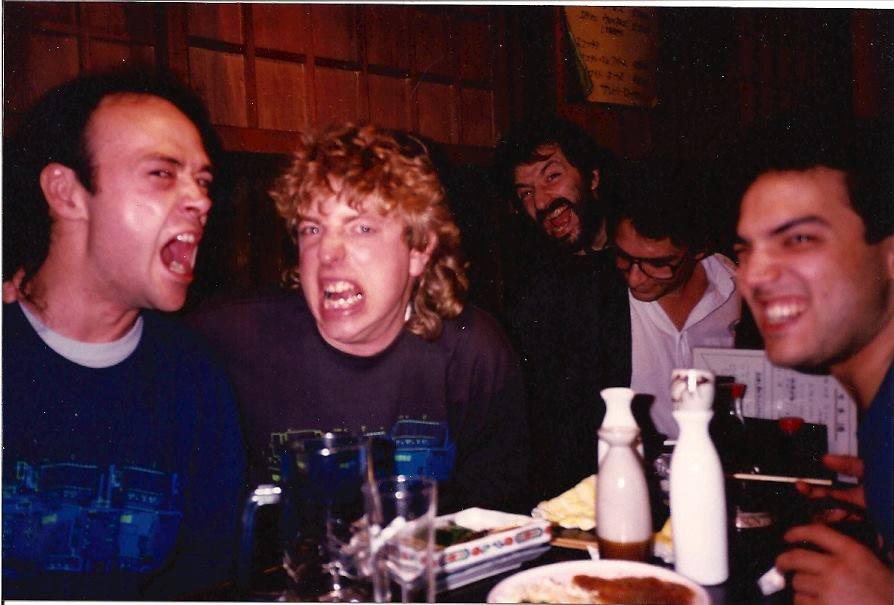Five men are sitting together at a table in a restaurant. Two men in front are making exaggerated facial expressions, while the others smile and laugh in the background. Plates, glasses, and bottles are on the table.