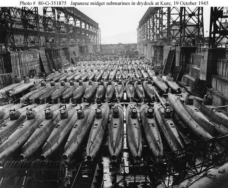 Rows of Japanese midget submarines sit tightly packed in a large drydock at Kure, Japan, surrounded by steel framework, with workers visible among the submarines. Photo dated October 19, 1945.