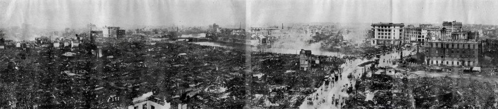 Panoramic black-and-white photo of a city devastated by fire; buildings are reduced to rubble, with a few structures partly standing, a river in the background, and people walking through the ruins.