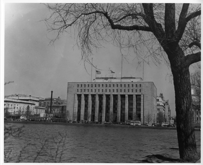 A large, rectangular building with tall columns stands beside a body of water, partially framed by a leafless tree in the foreground; several flags are flying on the building’s roof.