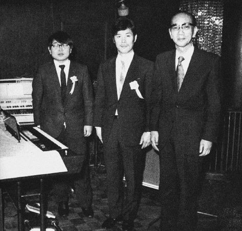 Three men in suits with ribbons stand indoors beside a keyboard instrument. The photo is in black and white and appears to be from a formal event or presentation.