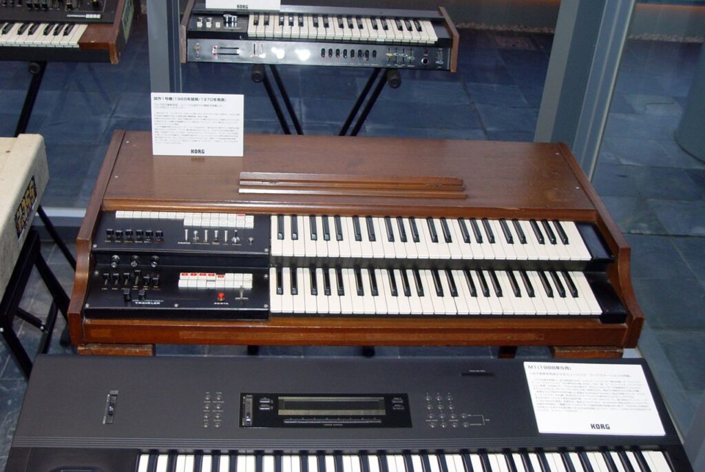A vintage Korg organ with two stacked keyboards and various control switches on the left sits on display, with informational placards and other keyboards visible in the background.