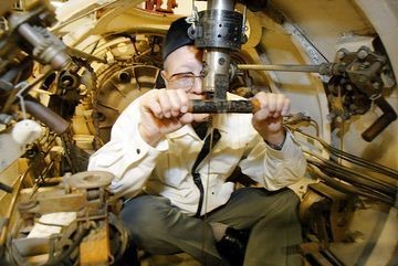 A person in a white shirt and dark hat operates machinery inside a confined, metallic space filled with mechanical components and pipes, possibly in a submarine or industrial setting.