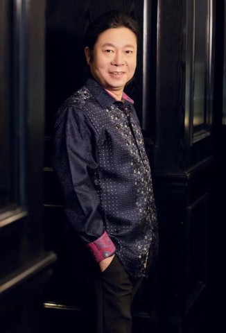 A man with dark hair wearing a dark patterned shirt with pink cuffs stands indoors against a dark wall, smiling gently at the camera.