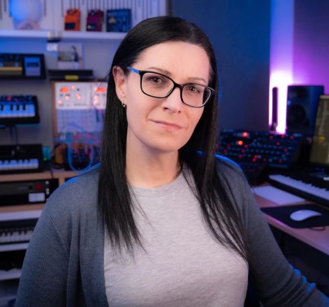 A woman with straight dark hair and glasses sits confidently in a modern music studio, surrounded by synthesizers, keyboards, and colorful audio equipment. The room is softly lit with blue and purple lights.
