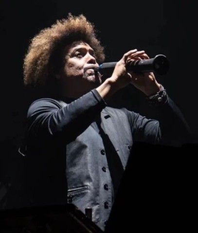 A man with an afro hairstyle wearing a dark jacket sings into a microphone on stage under dramatic lighting, with a dark background.