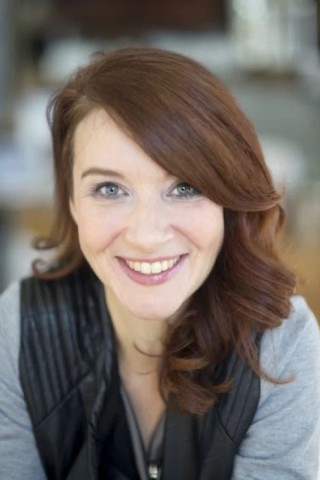 A woman with wavy auburn hair and light eyes smiles at the camera. She is wearing a gray top with a black vest. The background is softly blurred.