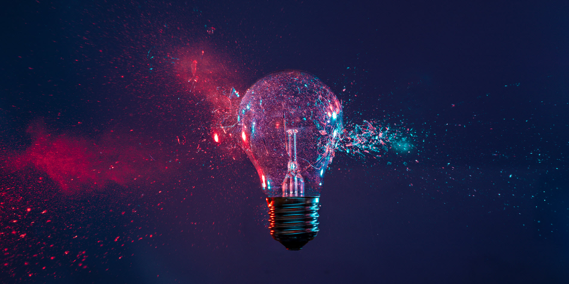 A light bulb shattering in mid-air, with red and blue particles and glass fragments exploding outward against a dark background.
