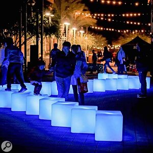 People walk and play on glowing blue light cubes in an outdoor area at night, surrounded by string lights, palm trees, and illuminated streetlights. The atmosphere appears lively and festive.