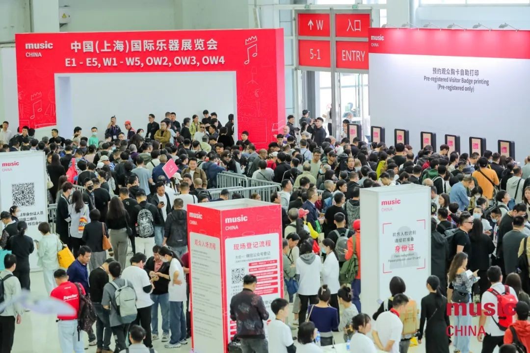 Large crowd of people gathers at the entrance of an exhibition hall for Music China in Shanghai; red and white signs display hall numbers and entry information in Chinese and English.