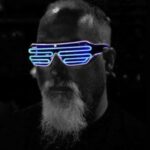 A man with a white beard is wearing futuristic, glowing blue LED glasses in a dark setting. His face is partially in shadow, highlighting the illuminated glasses.