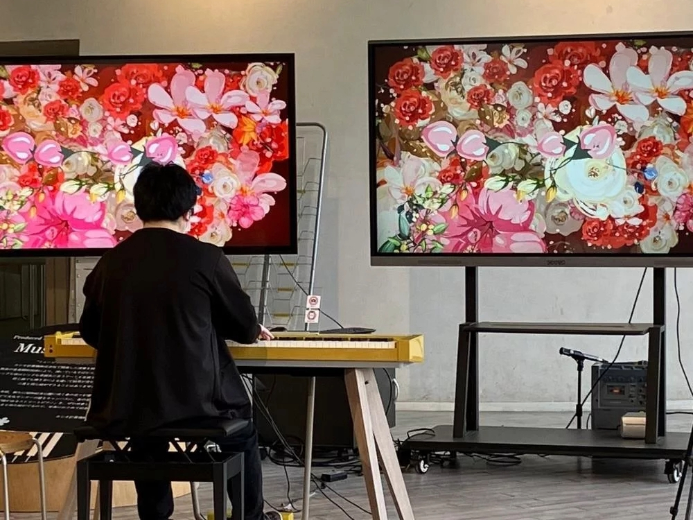 A person plays a yellow keyboard in front of two large screens displaying vibrant, colorful flower patterns. A microphone stand and audio equipment are nearby in a modern indoor setting.