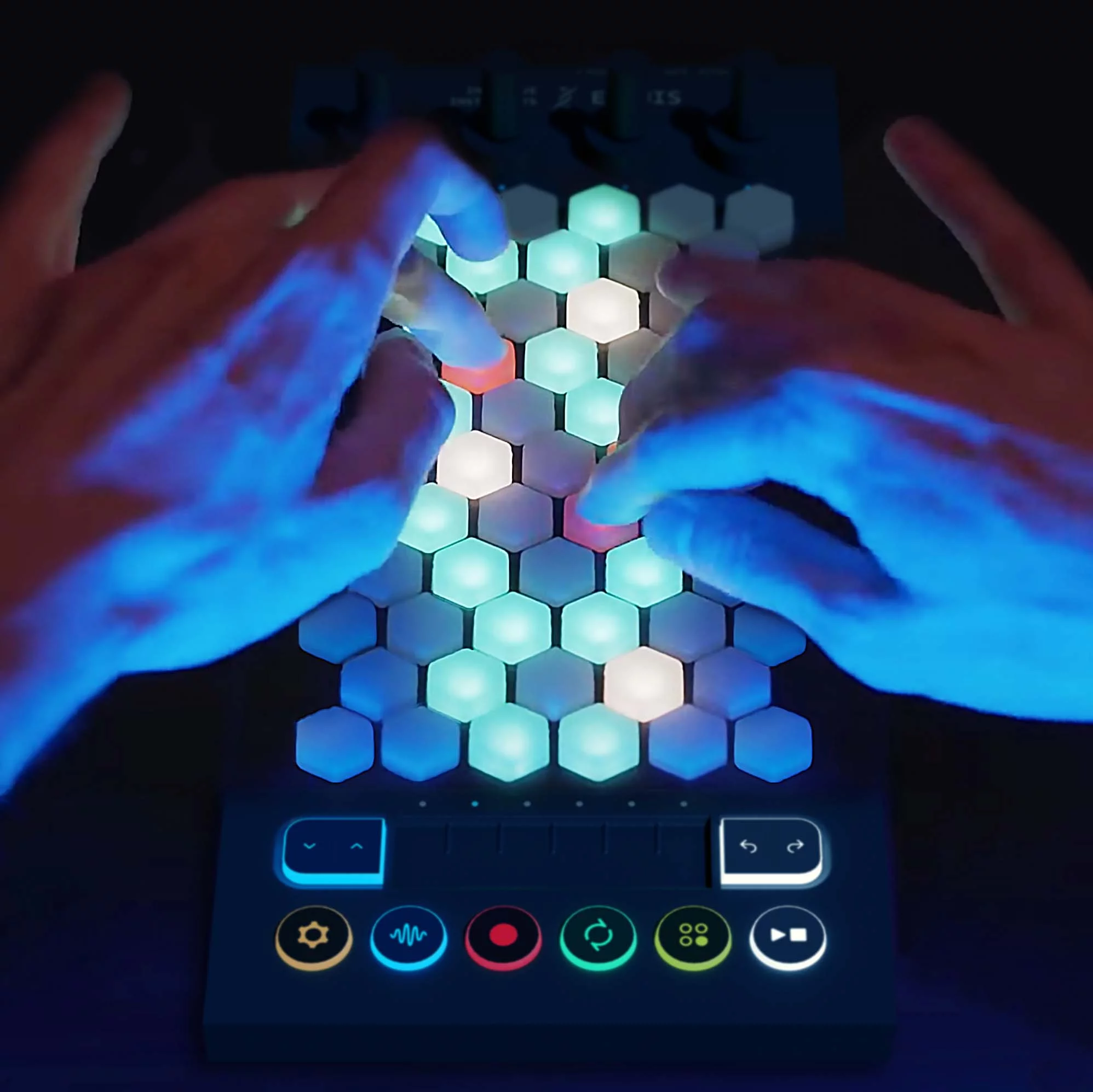 A pair of hands play a hexagon-shaped grid of illuminated buttons on an electronic music controller in a dark setting, with colorful icons and glowing lights.