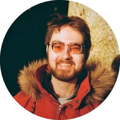 A person with short brown hair, a beard, and glasses is smiling while wearing a red parka with a fur-lined hood. The background is dark with a light-colored stone or wall behind them.