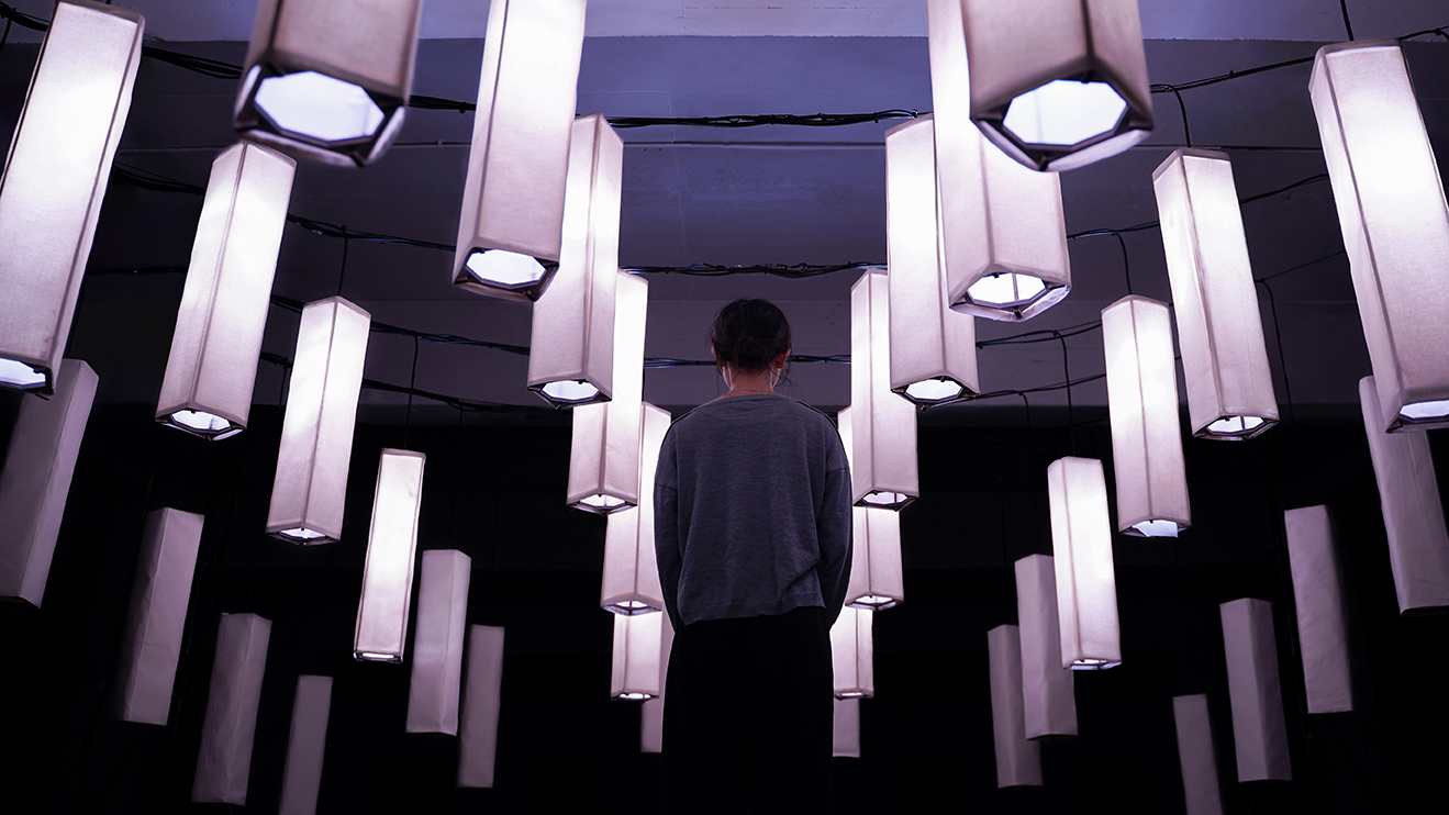 A person stands facing away, surrounded by many hanging rectangular lanterns with soft white light, creating an immersive and atmospheric indoor scene.