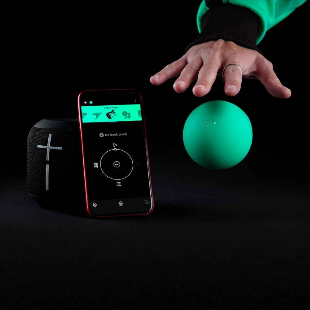 A hand hovers above a bright green ball next to a black cylindrical speaker with a plus sign and a smartphone displaying a music control app, all on a black surface with a dark background.