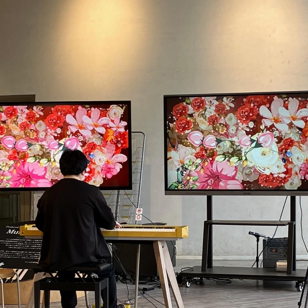 A person in black sits at a keyboard in front of two large screens displaying vibrant digital art with pink, red, and white flowers. The setting appears to be a modern indoor space with minimal decor.