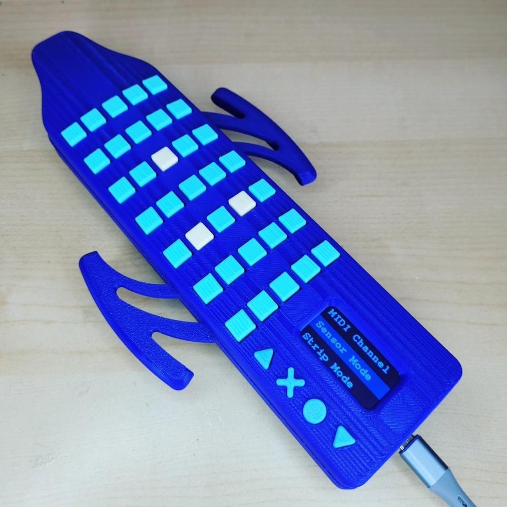 A blue electronic musical device with square buttons, curved side grips, and a small screen displaying MIDI settings, placed on a light wooden surface.