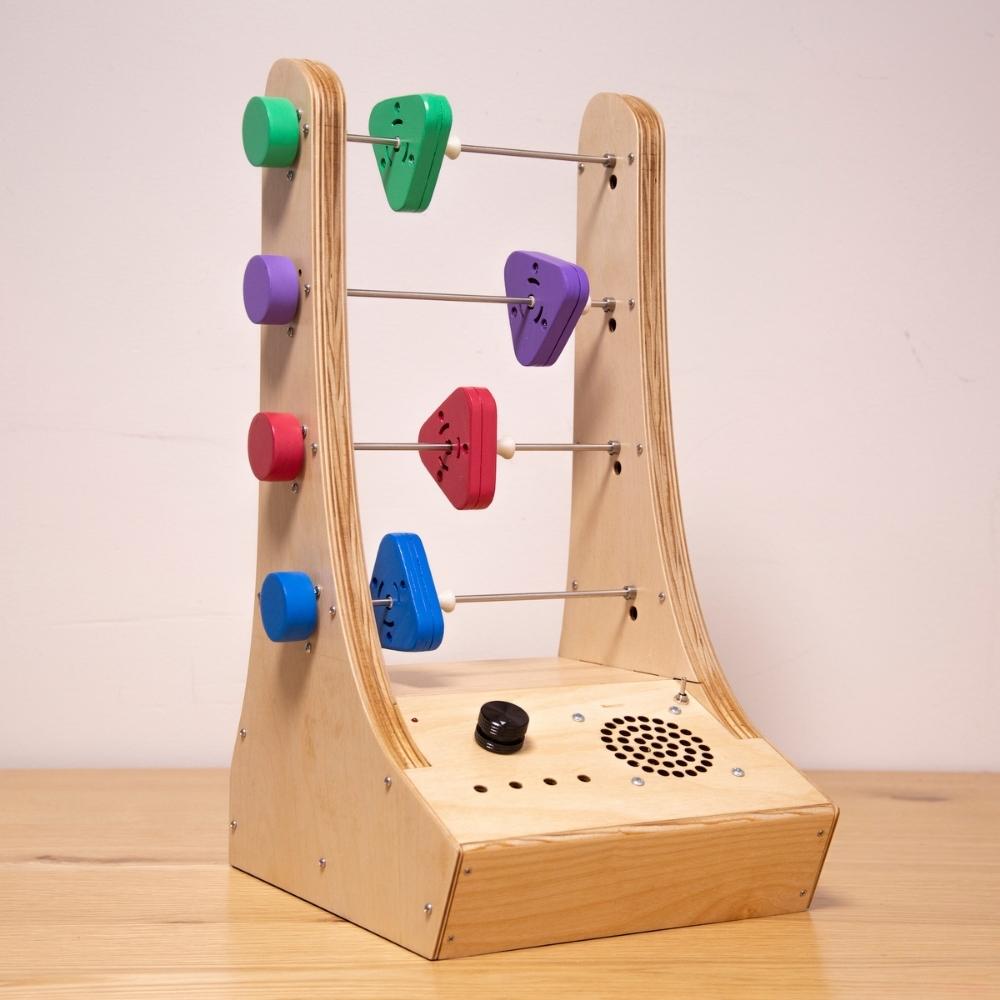 A wooden electronic music device with three metal rods, each holding two colorful sliders, and a speaker with control knobs on the front panel, placed on a light wooden surface.