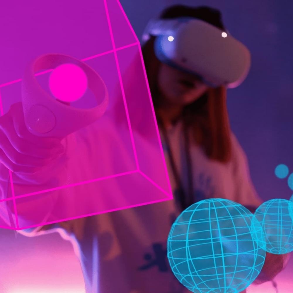 A person wearing a virtual reality headset interacts with floating neon pink and blue holographic shapes, including a cube and spheres, in a digitally immersive environment.