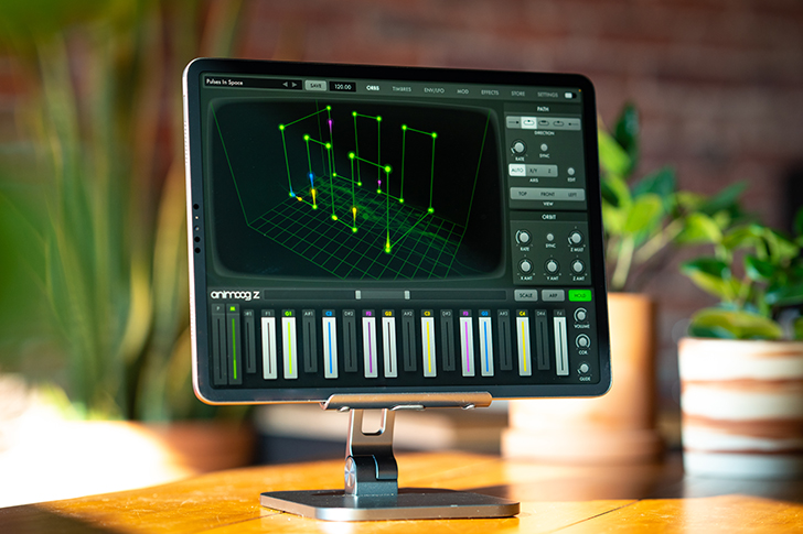 A tablet on a stand displays a music synthesizer interface with virtual keys and 3D modulation controls. The background is softly blurred, featuring green plants and warm lighting.